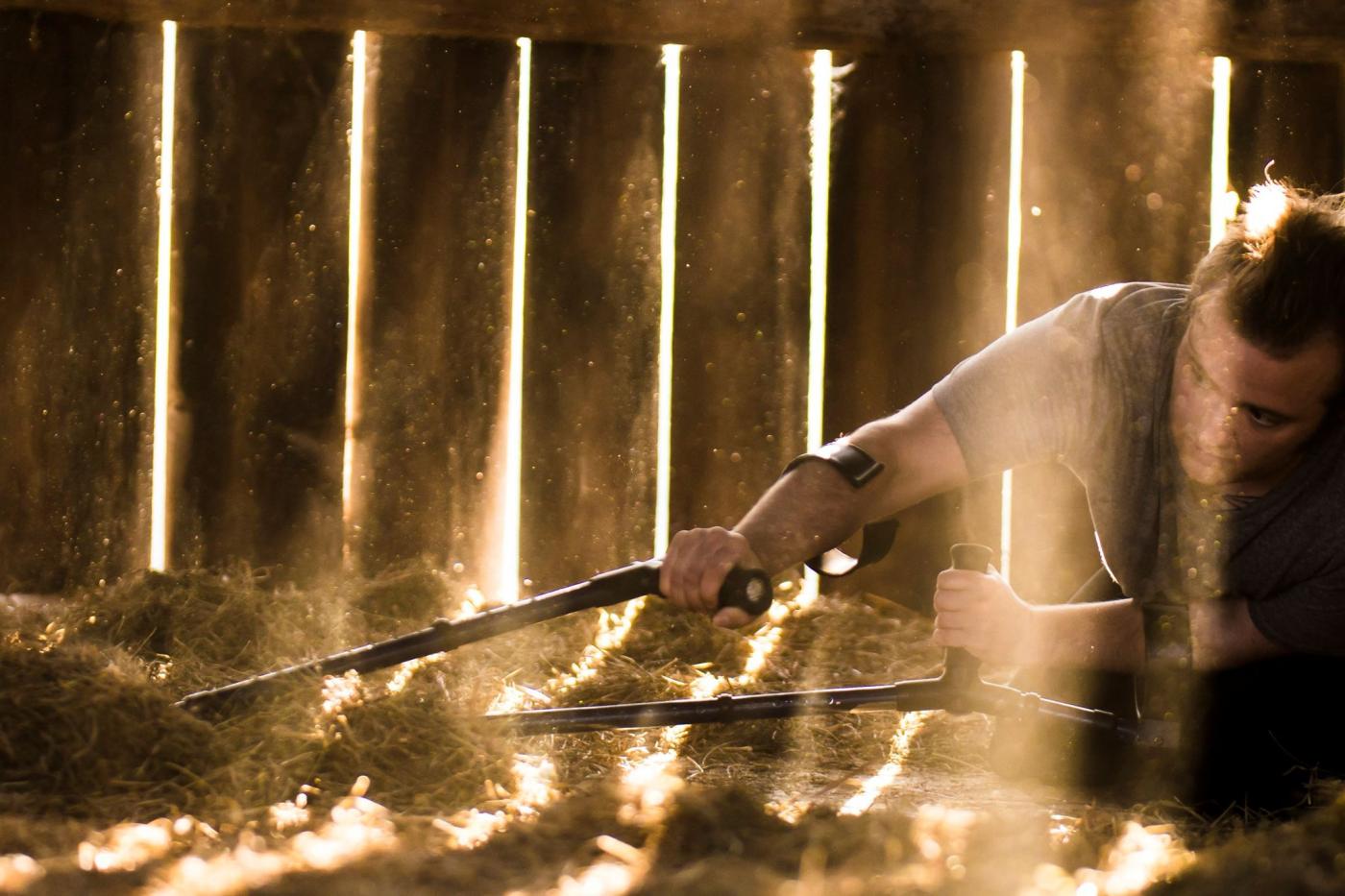 Toby lying on ground in a barn structure, pulling crutches through the hay in front of wooden slatted wall.