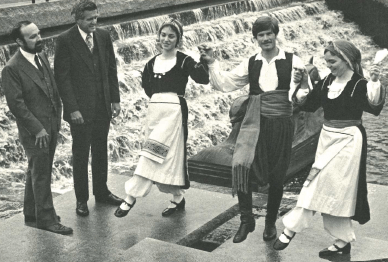 Two white men, in suits, watch three dancers in Swedish folk clothes by a waterfall in a city center.
