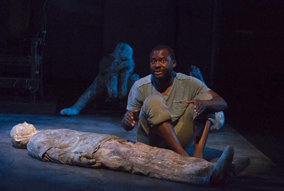 A man dressed in a t shirt, rolled up pants, and no shoes sits on a darkened stage. A prone figure is on the floor in front of him; other figures are in the background.