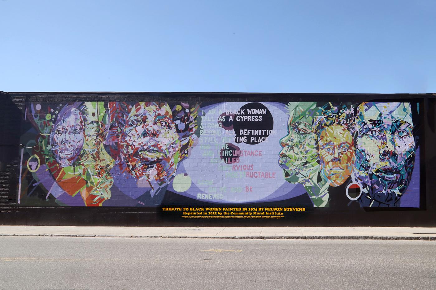 Outside on a sunny day, a mural (on a one story building) of Black women in greens, blues, oranges, and purples.