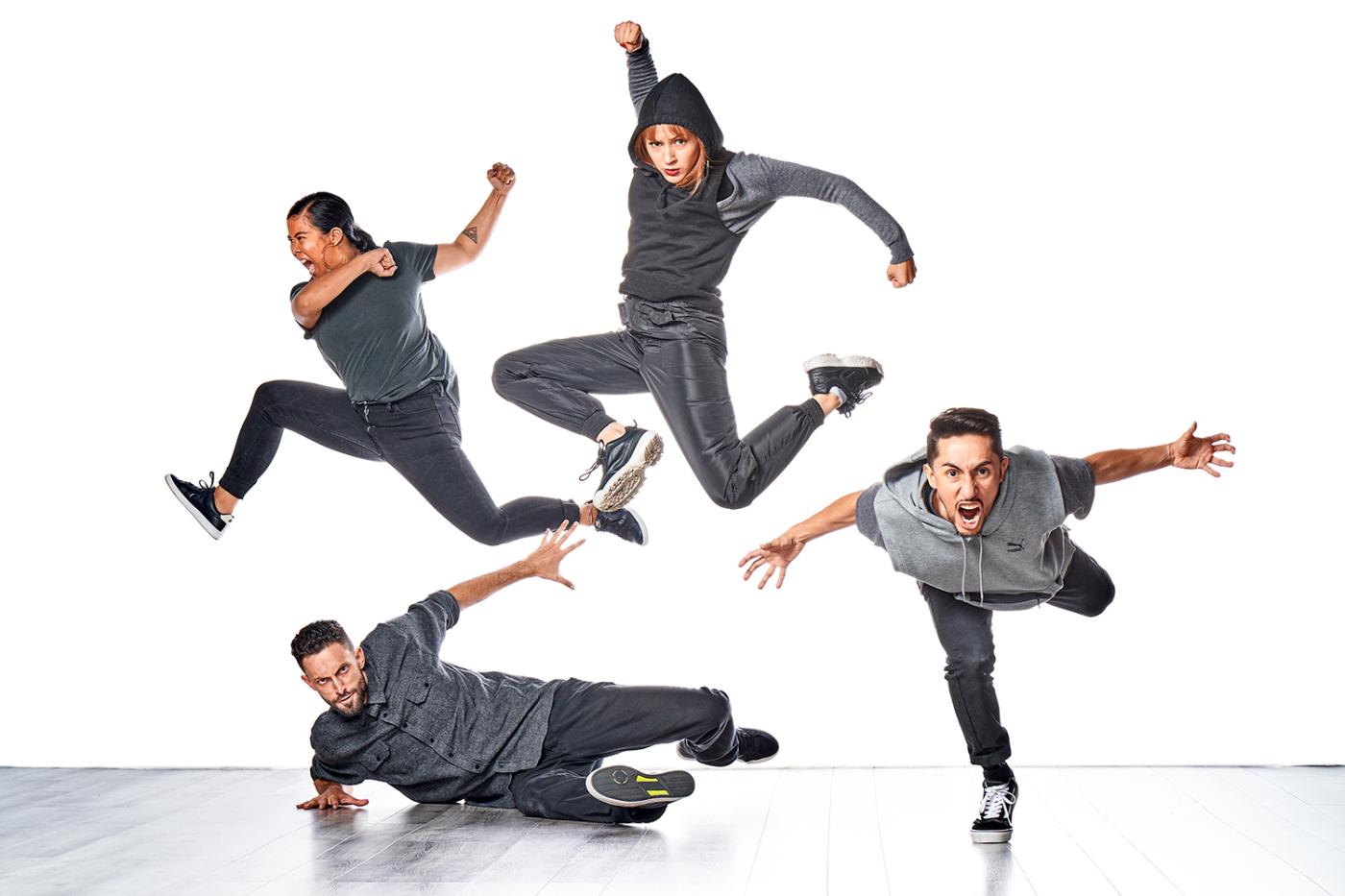 Four dancers, wearing gray, leap up, forward, or along the floor in front of a white backdrop.