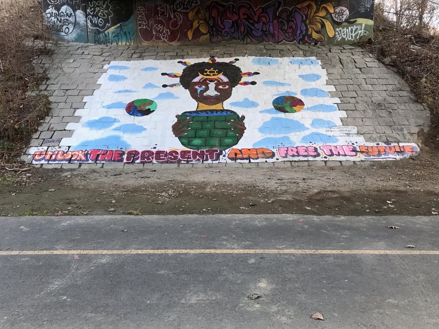 A large mural of a Black person with a shell like a turtle. "Unlock the present and free the future," it reads amongst blue clouds.