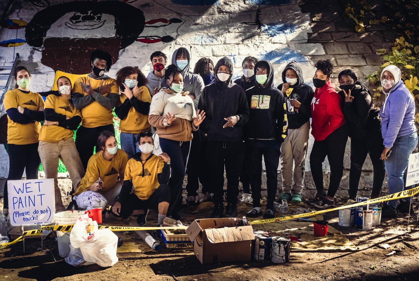 A large group of folks, half in yellow t-shirts, pose together in front of a large mural.
