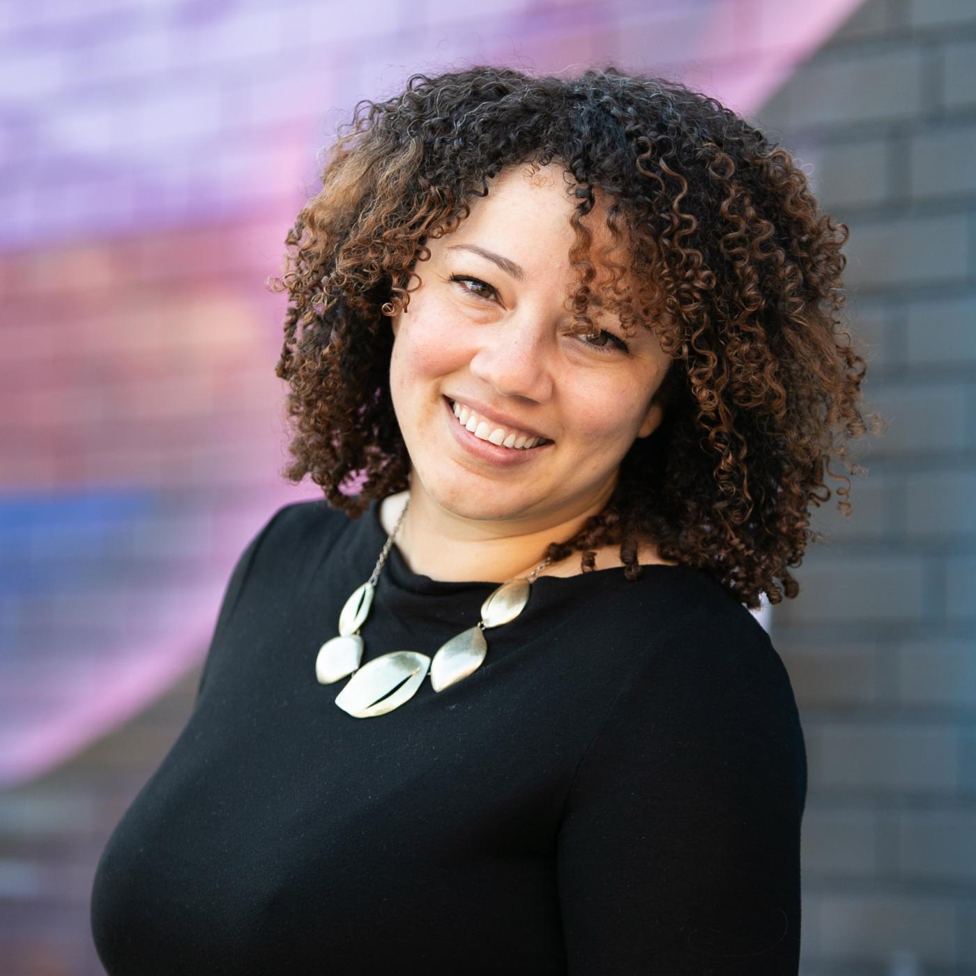 A lights-kinned woman smiles. She has shoulder length curls and wears a metallic necklace over a black blouse.