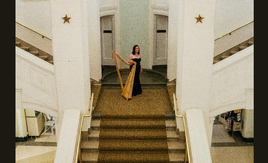 a woman poses with her harp at the top of a fancy staircase at an opera house