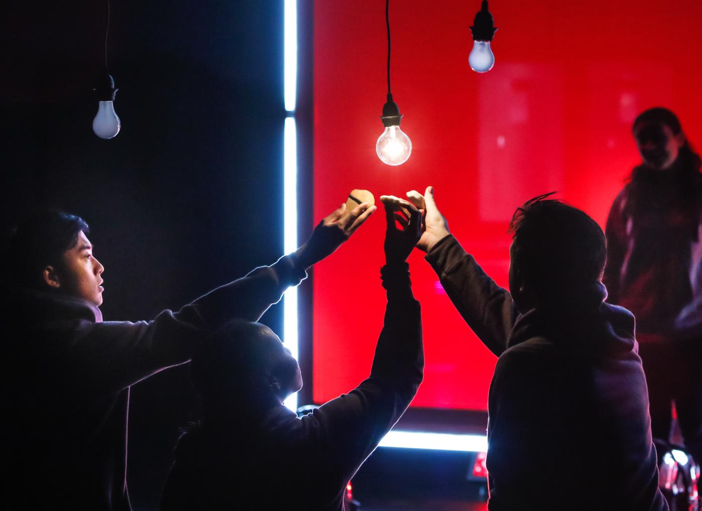 A group of dramatically lit performers all reach toward one lit light bulb, hanging on a wire from the ceiling.