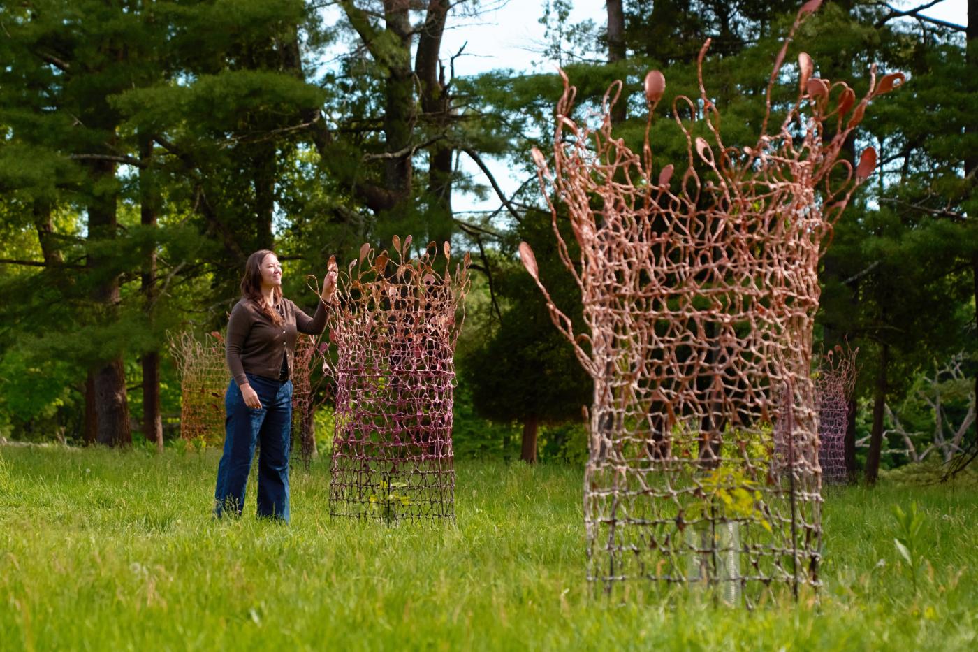In a grassy field, a woman, with dark brown hairs, stands next to three nets with plants that are about ten feet apart.