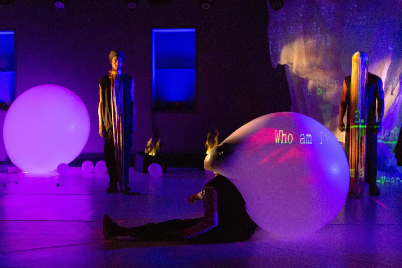 In a gallery, a dancer (with a mask that inflates like a balloon in the back) sits on the ground in front of another dancer who stands (and wears a long tassled mask).