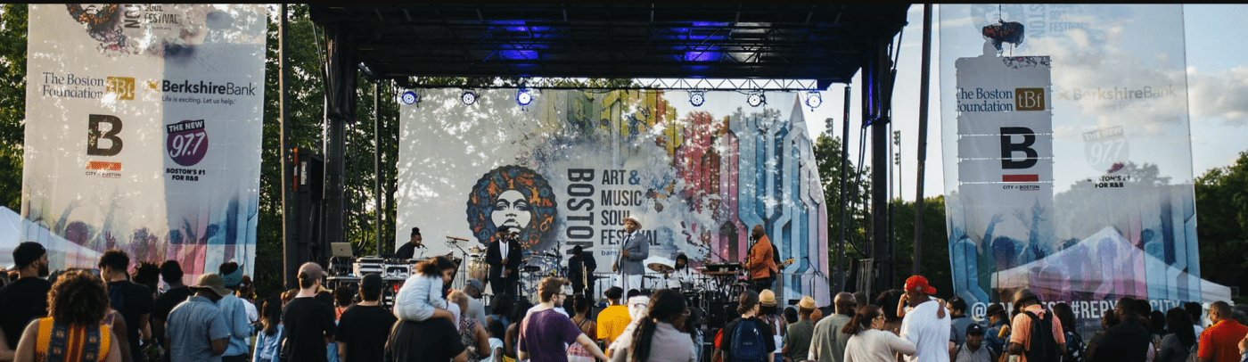 An outdoor stage on a sunny day with the BAMSfest logo on the screen; musicians are on the stage and there is a crowd gathered in front of the stage.