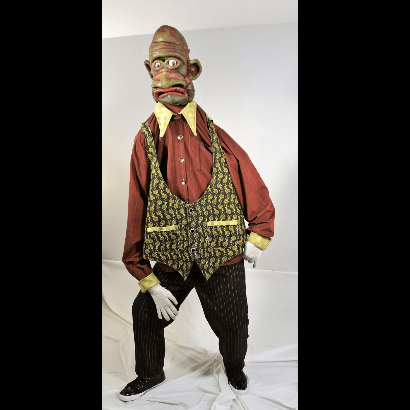 a giant grumpy looking puppet wears one of Leslie's creations: a green patterned vest over an orange, patterned, colalred shirt, with black dress pants.