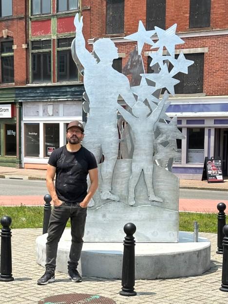 D Derek poses with the back of the steel sculpture, which features a larger man and a smaller boy with their arms stretch to the sky.