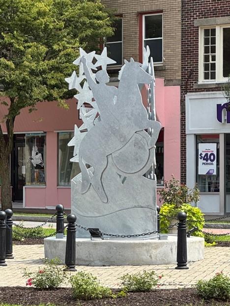 A steel sculpture of a cowboy riding a horse. Stars align the left side. It's in the middle of a town square.