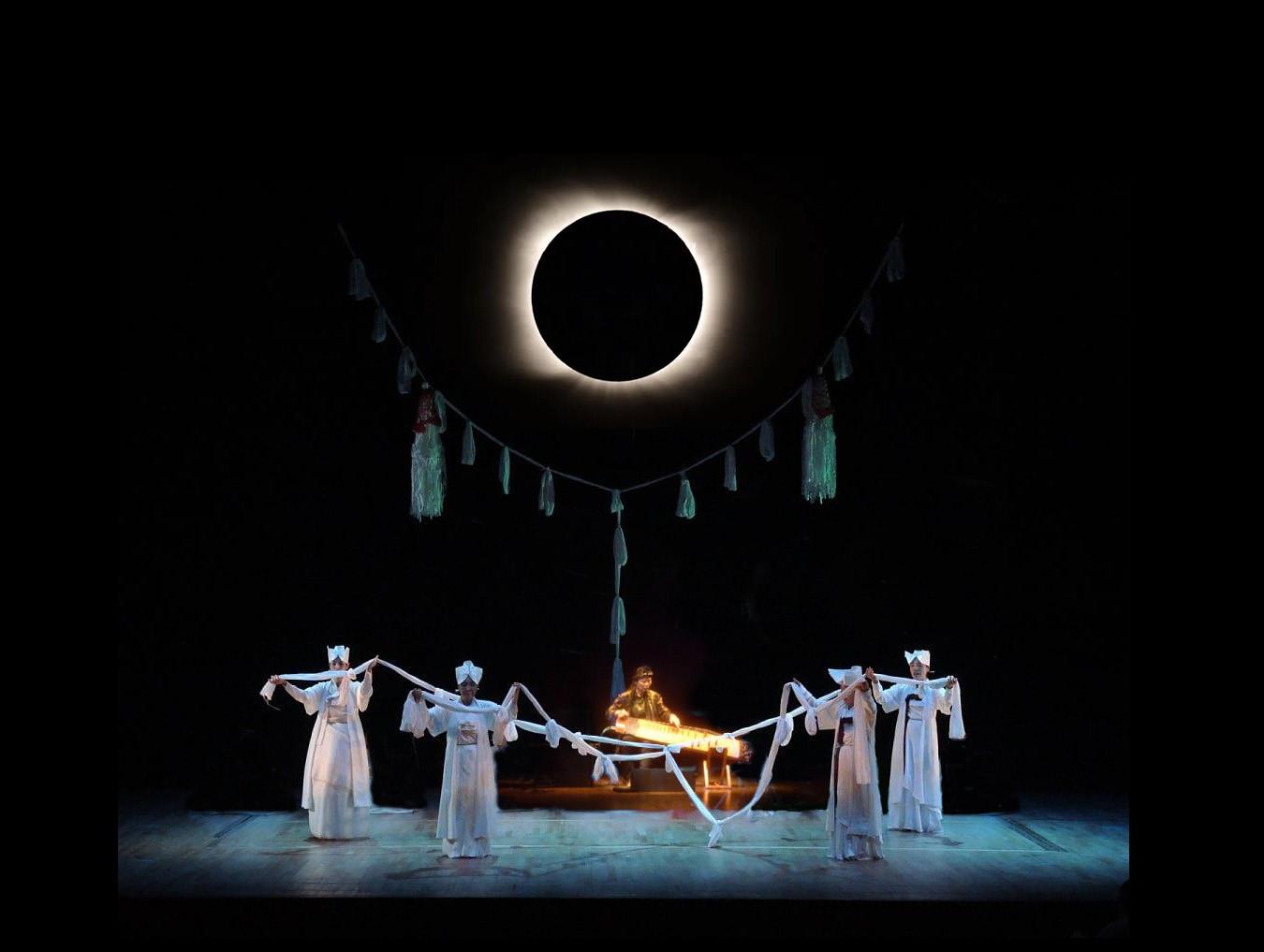 On dark stage, lit by an eclipse-like orb, four dancers in all white perform in front of Jin Hi who plays a string instrument behind them.