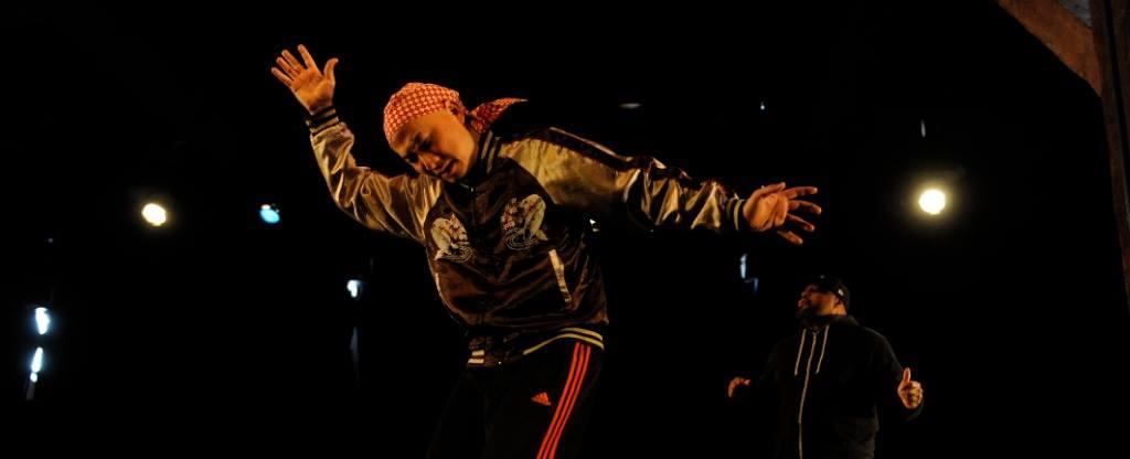 Two performers on a dimly lit stage; Michael is in front of Rennie performing a butoh-inspired movement while dressed in hip-hop attire.