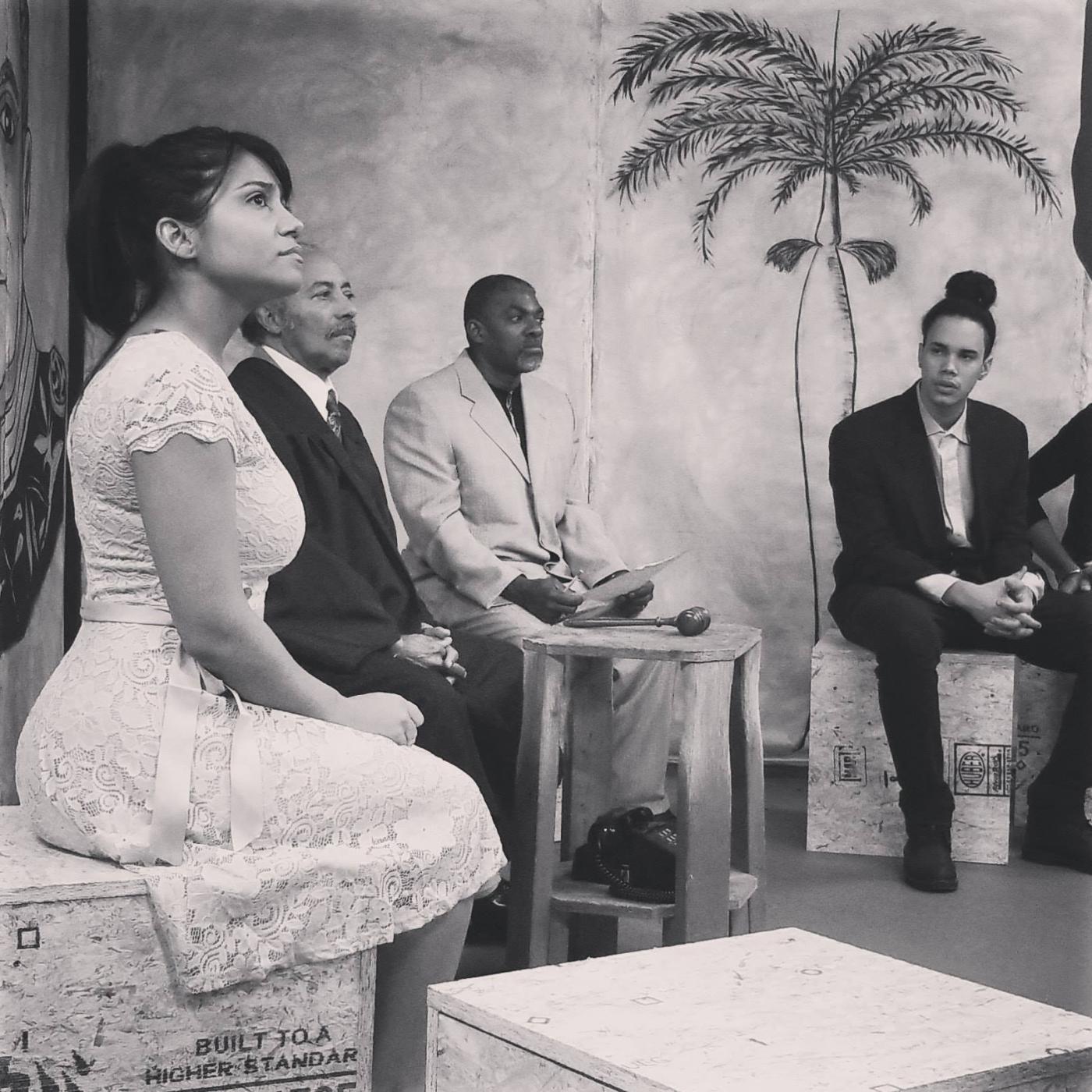 In black and white, a group of people sit in a lobby with a painted palm tree on the wall.