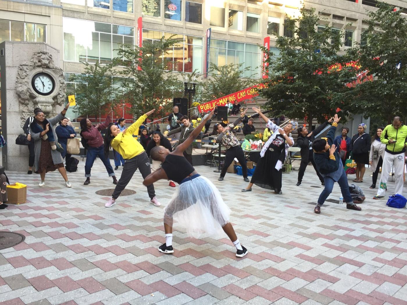 In an outdoor cobble-stone space, folks dance with their shoulders dipped and opposing arm in the air.
