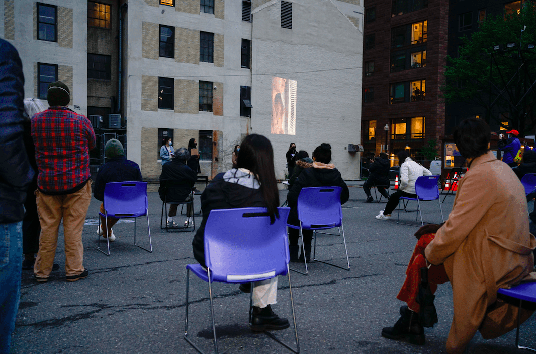 In a parking lot, folks watch a projection in blue chairs that are evenly spaced apart.