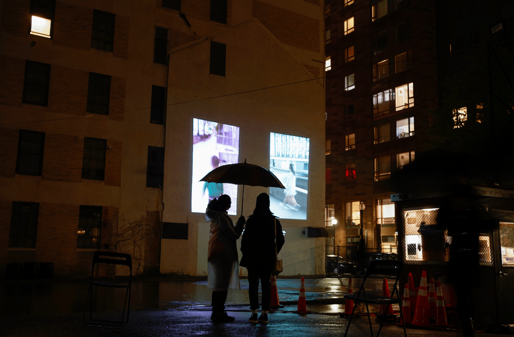 In a parking lot at night, a couple stands under an unbrella and watches projections on the side of a building.