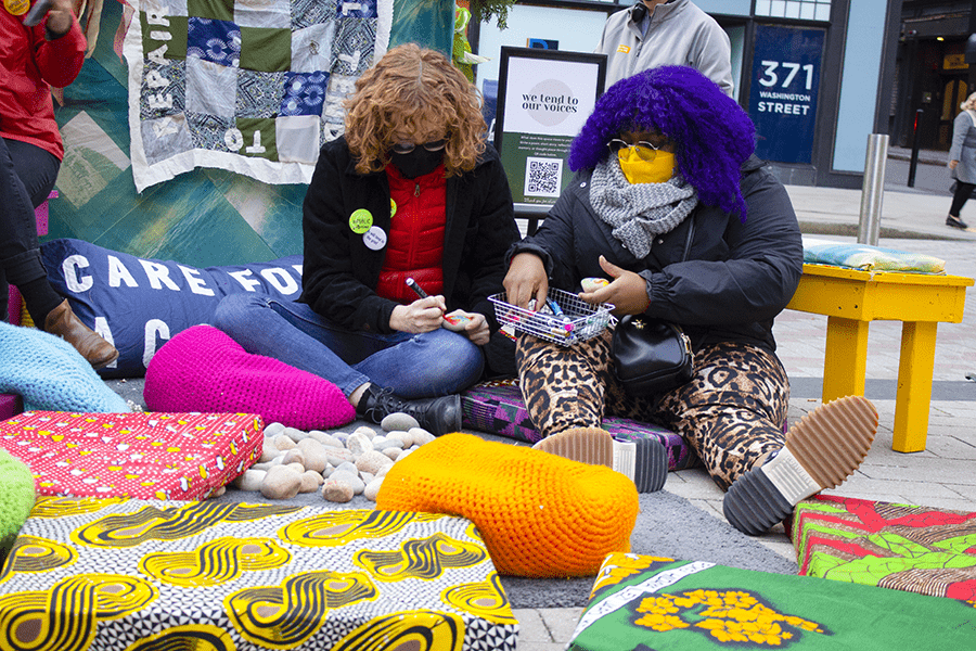 On the ground, two masked folks sit by some pillows. One of them has bright purple hair.