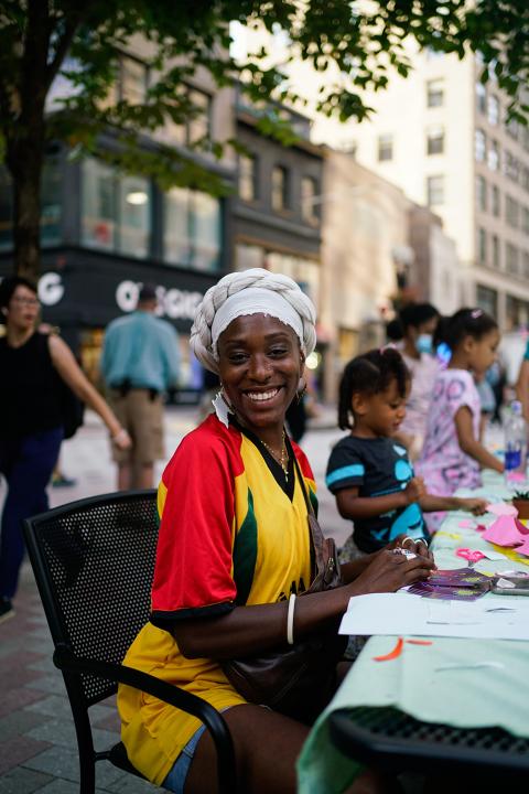 A Black woman, in a head wrap and bright red and yellow dress, smiles.