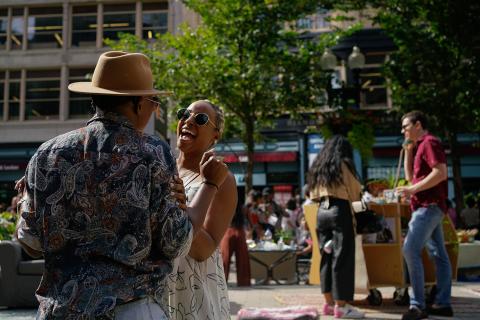 Two folks laugh together. One wears a big tan fedora. The other wears sunglasses.