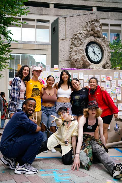 Nine folks pose together in front of a big clock and a wall of illustrations.