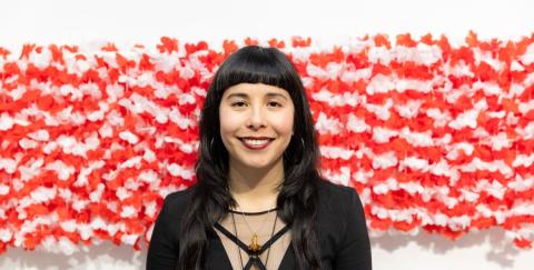 Lani poses in front of a red and white striped work of art. They have short brows and long dark hair. They wear a mesh and black top.