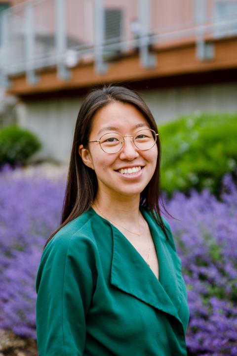 An Asian woman wearing a green blouse and glasses standing outside in a garden of purple flowers