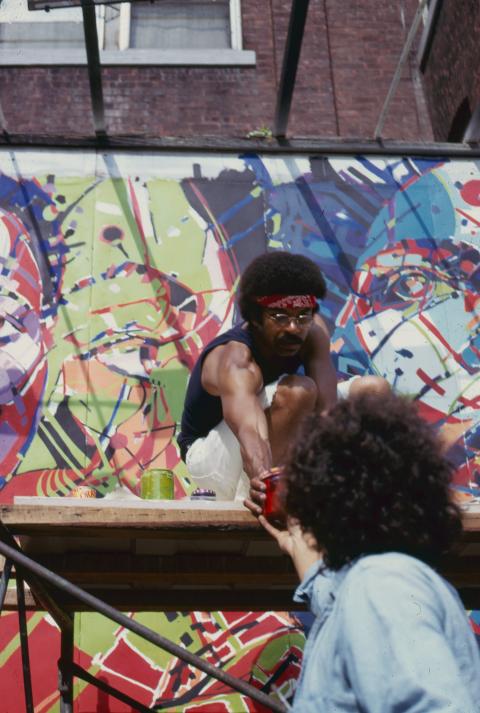 A Black man, with an Afro and in a navy tank top and cream short shorts, reaches for a can of paint from a friend who passes it to him.