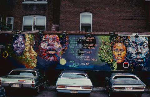 On a brick building, with cars parked (from behind), a mural of Black women in human and non-human, neon tones with text. Photo is scanned and aged.