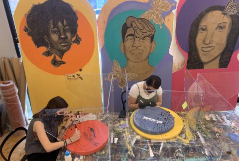 Two people in an art studio painting on round colorful discs. They are wearing masks and acrylic panels are placed between the. The background has 3 tall panels each with a colorful portrait of a person.