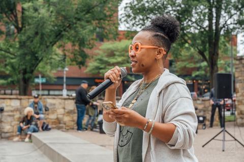 Ferene has her hair pulled back into a small 'fro. She is a Black woman. She speaks into a microphone and wears orange framed glasses.