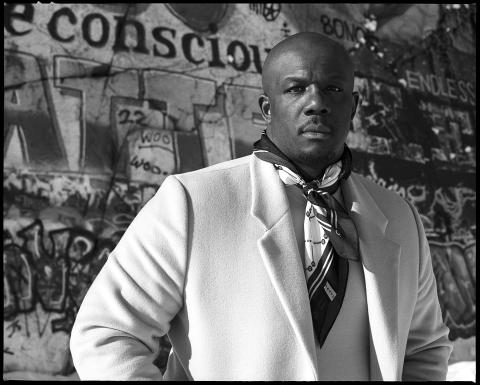 In black and white and in front of graffiti, a Black man poses in an ascot and white blazer.