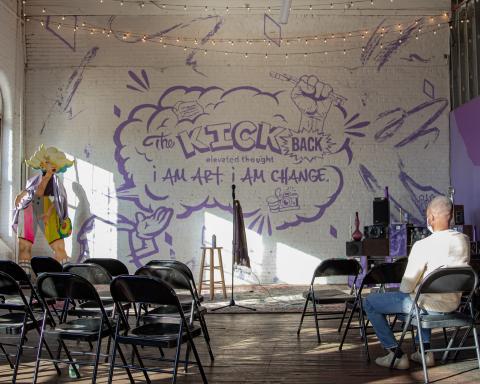 A performance space (microphone with chairs facing the microphone) with a mural behind it. A man sits in one of the chairs.