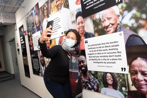 A woman, wearing a face mask, poses with a banner of images and quotes from folks in Lowell.