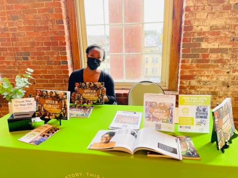 Christa sits behind a table. On the table is a book, with the title "Hidden in Plain Sight: Stories of Black Lowell."
