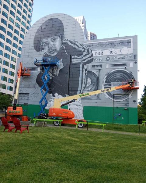 Forklifts in front of an in-progress mural of a young Black boy posing in front of a boombox with a purple cassette tape inside it.