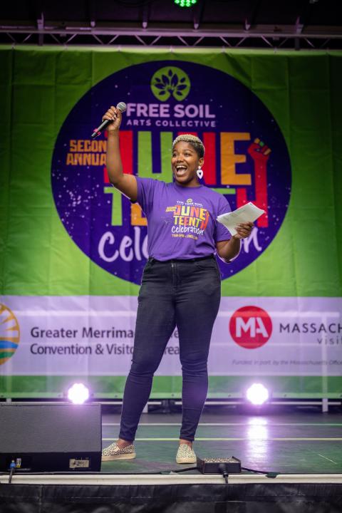 On an outdoor stage, Christa holds a mic and wears a purple t-shirt that matches the sign behind her.