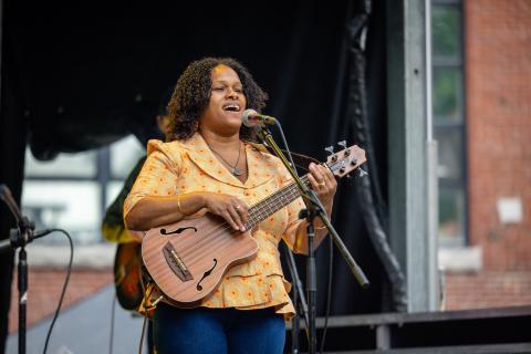 On an outdoor stage, a woman of color plays the guitar and sings into a microphone.