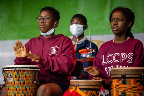 Three kids, wearing maroon shirts and face masks, play the bongos.
