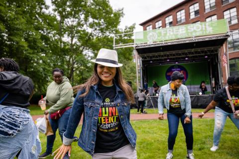 In a field, women of color, in event t-shirts, dance together. One of them, in the foreground, wears a fedora and smiles.