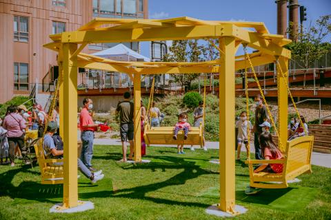 A yellow hexigonal swing set has three swings all facing each other. Folks sit on it.