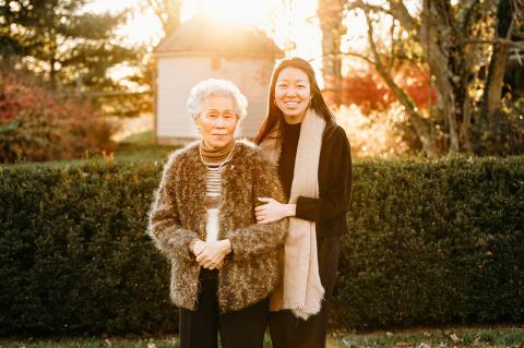 An elderly Asian woman and a young Asian woman pose together. The elderly woman wears a fuzzy sweater and the young woman wears a tan scarf. They're outside by some shrubs at sunset.