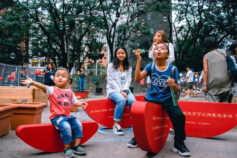 Three Asian kids blow bubbles and sit on a red circular public art installation.