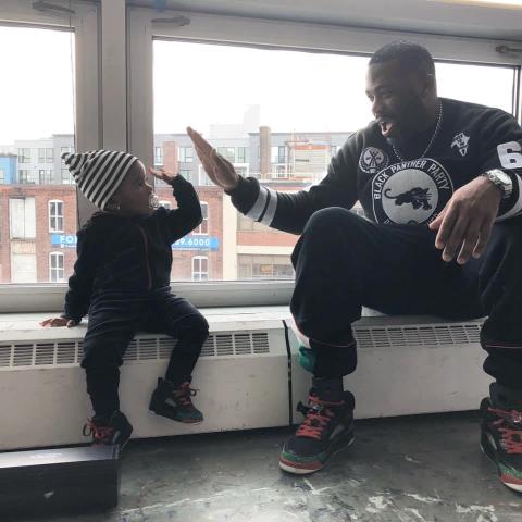 A Black man, wearing all black, high fives a young Black girl, in a white beanie and all black clothes.