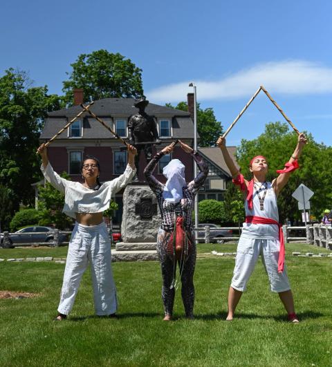 Three folks stand in front of a monument of a white man. These folks hold arches over their heads and are wrapped in ribbons. One of the three wears the white shroud. 