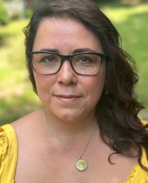 A woman with long dark hair, glasses, a gold pendent and a yellow blouse.