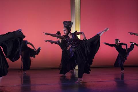 A dancer, in an ensemble of dancers, lifts her leg back and up and points forward.