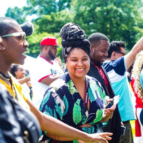 On a sunny day and in a crowd of dancing folks, a woman, with her braids in a bun and a floral print dress, smiles and dances.