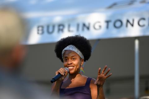 A Black woman, with a short afro, speaks into a microphone. Behind her a sign reads "Burlington."